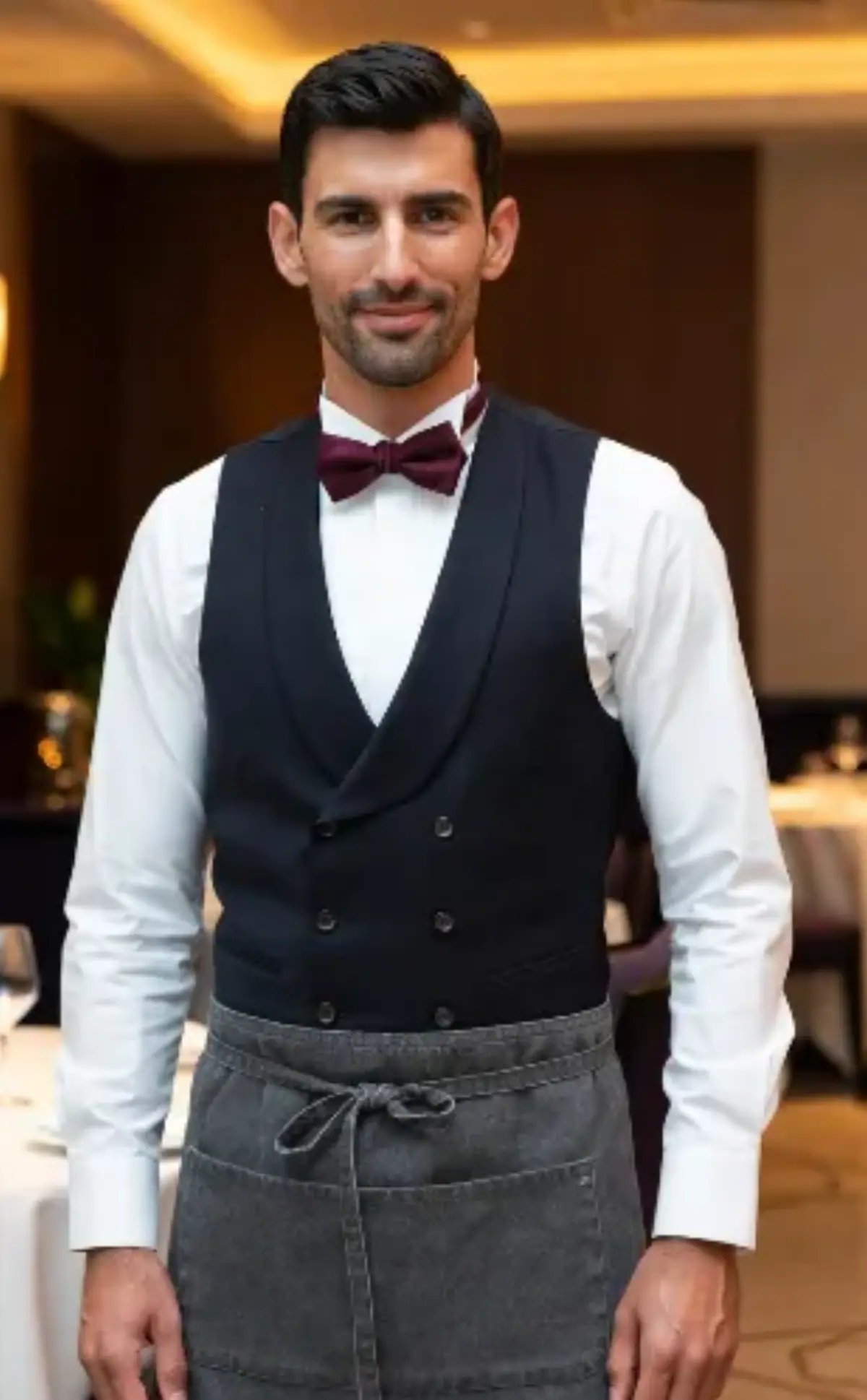 Un uomo di circa 30-35 anni, carnagione olivastra, capelli castani corti e ben pettinato, barba curata corta. Sorriso cordiale e professionale, sguardo diretto in camera che trasmette fiducia e accoglienza. Uniforme classica da Chef de Rang ristoranti rurale di livello