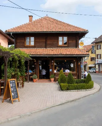 Un ristorante tradizionale in un borgo storico, con facciata in legno scuro e tetto di tegole rosse. All’ingresso una pergola coperta di edera e rampicanti, tavoli esterni su pavimentazione in pietra con vasi di arbusti curati. Un cavalletto con lavagna del menu è posizionato sul marciapiede, mentre all’interno si vedono clienti e personale. Sullo sfondo altre case tipiche del borgo, una strada tranquilla con auto parcheggiate e cielo azzurro con poche nuvole. L’immagine comunica calore, autenticità e l’opportunità di trasformare un contesto storico in un business di successo attraverso la ristorazione locale.