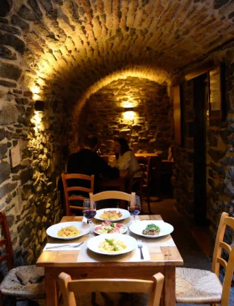 Interno rustico di una trattoria in borgo antico, soffitto a volta con archi di pietra grezza e travi di legno scuro. Luce calda da lampade a sospensione e candele. Al centro un tavolo di legno massiccio con esattamente 2 persone sedute una di fronte all'altra (una donna con capelli scuri in maglione rosso, l'altra con capelli chiari raccolti e grembiule chiaro). Sul tavolo 4 piatti visibili: Pasta con ceci (lagane e ceci, tonnellata di legumi e pomodoro) Cavatelli con peperoni cruschi e salsiccia Antipasto con salumi, formaggi pecorini e olive Secondo semplice (agnello alla brace con erbe) Due bicchieri di vino rosso, forchette, tovaglioli di stoffa, atmosfera intima e accogliente, niente secondo tavolo visibile, niente folla o altre persone in sfondo. Pareti in pietra con nicchie e bottiglie di vino.