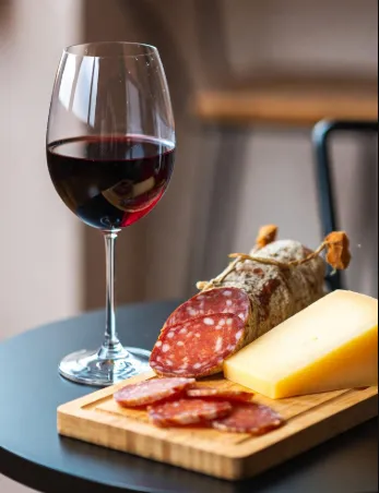 Calice di vino bianco (o rosato molto chiaro) riempito per metà, vetro trasparente con stelo lungo e calice ampio tipo ballon Tagliere di legno rettangolare con manico, appoggiato sul tavolo Sopra il tagliere: coltellino con lama nera e manico metallico Fette di salame con pepe in grani e puntini bianchi (tipo salame nostrano o felino-style) Pezzo di formaggio duro giallo intenso (tipo parmigiano o grana, ma tagliato a blocco) Alcune fette di salame arrotolate a fiorellino per decorazione