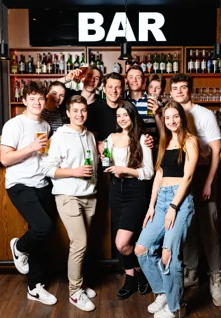 Gruppo di giovani europei chiari, sorridenti e abbracciati stretti davanti al bancone in legno scuro di un pub. Birre in mano (pinte e bottiglie), look casual (camice a quadri, jeans strappati, top neri), sfondo con spillatori birra, luci soffuse e insegna "BAR" illuminata, vibe amichevole e spensierata da serata tra amici.