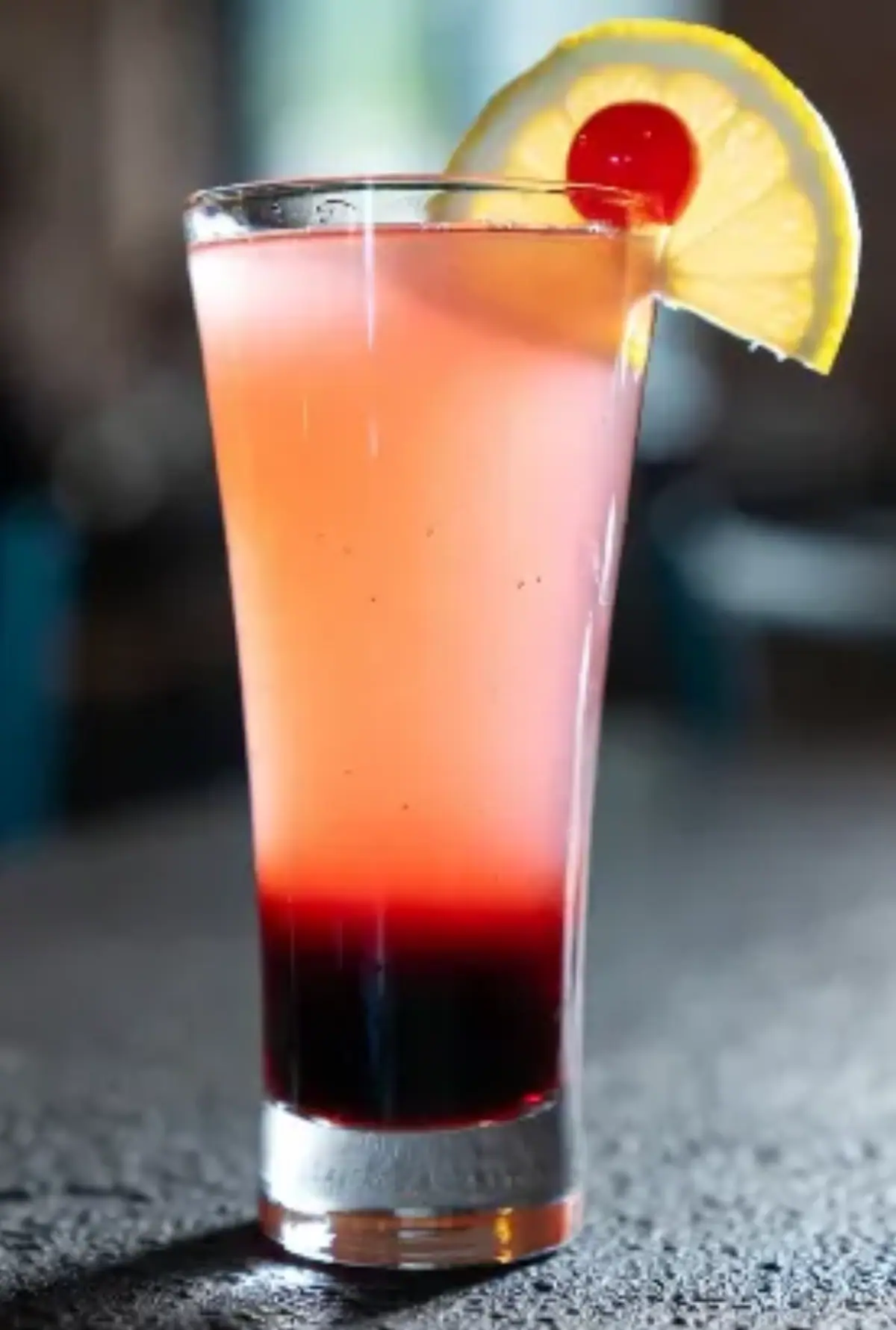Bicchiere: Alto e conico (tipo highball o flute slanciata), trasparente, con condensa visibile sulle pareti che suggerisce freschezza. Cocktail: Stratificato (layered) in tre colori graduali - fondo rosso scuro intenso (probabilmente grenadina o frutto rosso), parte centrale rosa chiaro opaco, superficie arancione-rosa sfumato con bollicine leggere. Guarnizione: Una fetta di limone giallo fresca sul bordo del bicchiere, con una ciliegia maraschino rossa al centro infilzata su uno stecchino. Sfondo: Sfocato in stile bokeh, con ambiente bar/ristorante caldo - luci soffuse, tavolo scuro in primo piano, figure indistinte e pareti in mattoni o legno. Sembra ispirata a un classico come "Tequila Sunrise" rivisitato o un "Sex on the Beach" variato,