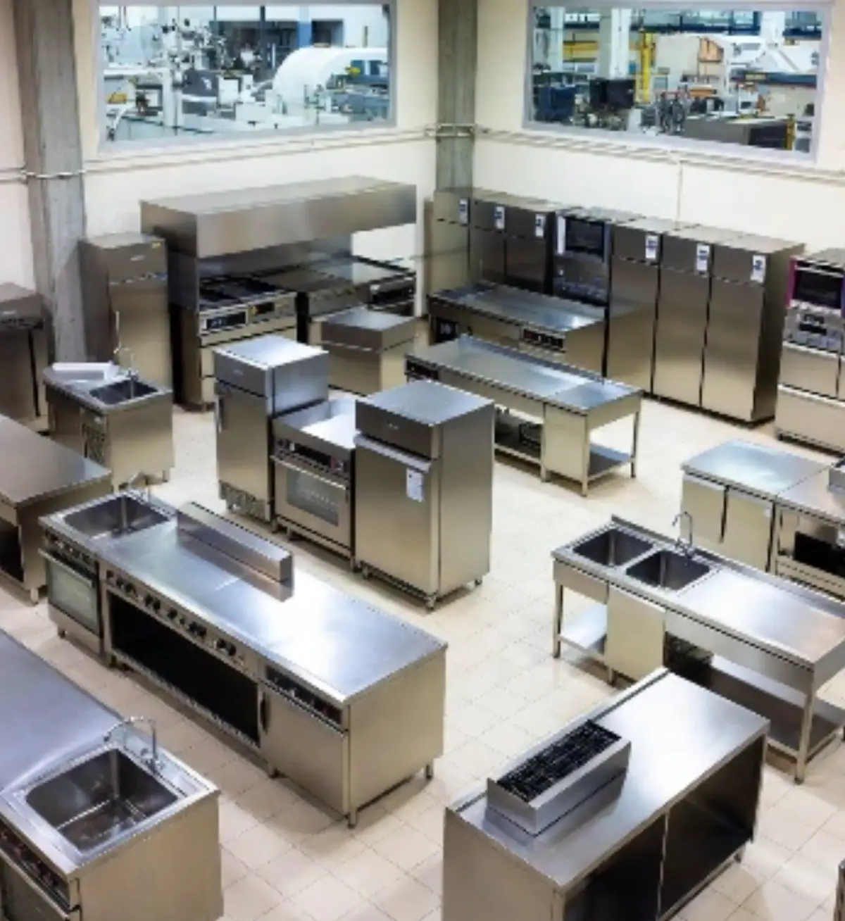 Una vista aerea dall'alto di un ampio showroom italiano moderno e luminoso, con pavimenti piastrellati chiari e illuminazione industriale. Al centro, sono esposte diverse cucine industriali in acciaio inox lucido: piani di lavoro modulari con fornelli integrati, lavelli doppi, forni a convezione, cappe aspiranti, tavoli refrigerati e armadi frigo.