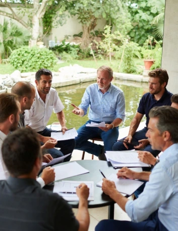 Un incontro più ristretto tra produttori agricoli e responsabili della distribuzione, seduti intorno a un tavolo rotondo in un patio aperto con vista su un giardino verde e uno stagno. Sul tavolo documenti, grafici colorati e fogli con dati. Un agricoltore anziano con camicia chiara sta spiegando qualcosa indicando i fogli, mentre gli altri ascoltano attentamente. L’atmosfera è professionale ma immersa nella natura, a sottolineare il tema della sostenibilità e del confronto tra canali distributivi tradizionali e vendita diretta.