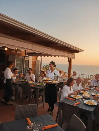 Terrazza panoramica di un ristorante sul mare al tramonto con 9 clienti seduti ai tavoli e 2 cameriere che servono piatti. Atmosfera calda e conviviale con vista mozzafiato sul mare.