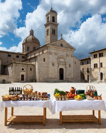 In primo piano due tavoli in piazza lunghi coperti da tovaglie bianche: sul tavolo di sinistra ci sono cesti di pane, pagnotte, bottiglie di vino rosso, barattoli di miele e confetture; sul tavolo di destra verdure fresche (zucchine, pomodori, peperoni), limoni, brocche di terracotta e olio. Sullo sfondo domina una maestosa cattedrale barocca con facciata classica, colonne, cupola e campanile alto, stile tipico italiano
