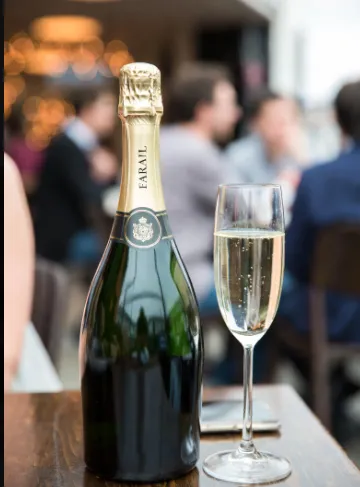 Bottiglia verde scuro in primo piano su tavolo di legno scuro. Capsula dorata, etichetta nera con scritta "FARAL" in bianco e piccolo stemma centrale (anche questo puramente decorativo, generato casualmente). Davanti alla bottiglia, un flute con vino chiaro, bollicine visibili e riflessi luminosi. Sul tavolo si intravede un telefono sfocato. Sfondo: persone sedute e in piedi in un ambiente luminoso (pare un bar o terrazza coperta), luci bokeh calde, atmosfera rilassata da aperitivo. Tonalità più naturali, luce diurna/serale mista.