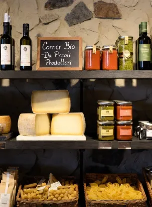 “Corner Bio” all’interno di un affascinante negozio gourmet italiano di alta gamma. Illuminazione naturale calda e morbida che crea un’atmosfera accogliente e di lusso. Bellissime mensole in legno scuro pregiato e parete in pietra naturale grezza. I prodotti biologici artigianali sono disposti con cura ed eleganza: eleganti bottiglie di olio extravergine di oliva (alcune beige, altre verdi), grandi forme di formaggi stagionati su piani di marmo bianco, barattoli di vetro con salse pregiate (pomodoro e pesto), vasetti di miele, prodotti al tartufo e pasta secca artigianale di lusso in cestini di vimini. Al centro un piccolo cartello in gesso dal gusto raffinato con la scritta “Corner Bio – Da Piccoli Produttori”.