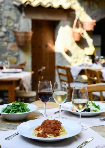 Interno realistico di una vecchia osteria in borgo di pietra. Tavolo di legno grezzo con tovaglia bianca semplice. Al centro un piatto di spaghetti al ragù con carne macinata e pomodoro, sopra una foglia di basilico fresco e un po’ di parmigiano grattugiato. Accanto: ciotola di insalata di rucola e pomodorini, due bicchieri di vino (uno rosso, uno bianco), forchetta e coltello posati, luce calda da una lampada appesa al soffitto con travi di legno scuro. Sfondo: muro in pietra grezza, porta antica socchiusa che dà su un vicolo, qualche bottiglia di vino sullo scaffale. Atmosfera intima, vissuta, niente turisti o roba patinata.