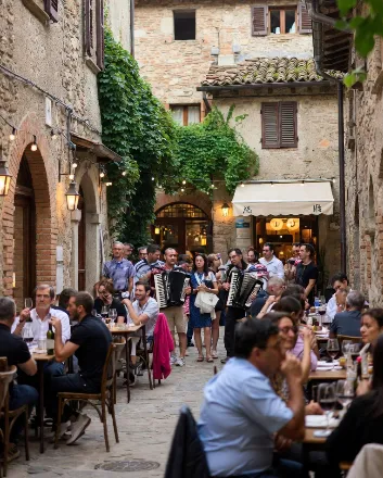 Vicolo stretto di un borgo medievale italiano, pavimentato in pietra irregolare, con muri in mattoni antichi e finestre con persiane marroni. Al centro, due musicisti (uomini con camicia bianca e pantaloni scuri) suonano la fisarmonica, uno con strumento rosso e nero, l’altro classico nero; sorridono e coinvolgono il pubblico. Intorno, tavolini all’aperto con sedie di legno: gente seduta che beve vino rosso e bianco da calici, mangia piatti semplici (pane, formaggi, salumi). Turisti internazionali evidenti: gruppi di asiatici (cinesi o orientali, con borse e macchine fotografiche), americani (stile casual con polo e cappellini), europei misti; alcuni in piedi che fotografano o applaudono i musicisti.