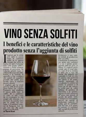 pagina di giornale, pulita e centrata, con titolo -VINO SENZA SOLFITI- in nero bold maiuscolo. Sottotitolo lungo: "I benefici e le caratteristiche del vino prodotto senza l’aggiunta di solfiti". Al centro una foto grande di un calice di vino rosso su tavolo di legno, Il testo dell’articolo circonda la foto, con un occhiello iniziale evidenziato e corpo in colonne. Firma "di Maria Rossi" in basso a sinistra. inserto enogastronomici di quotidiani nazionali.