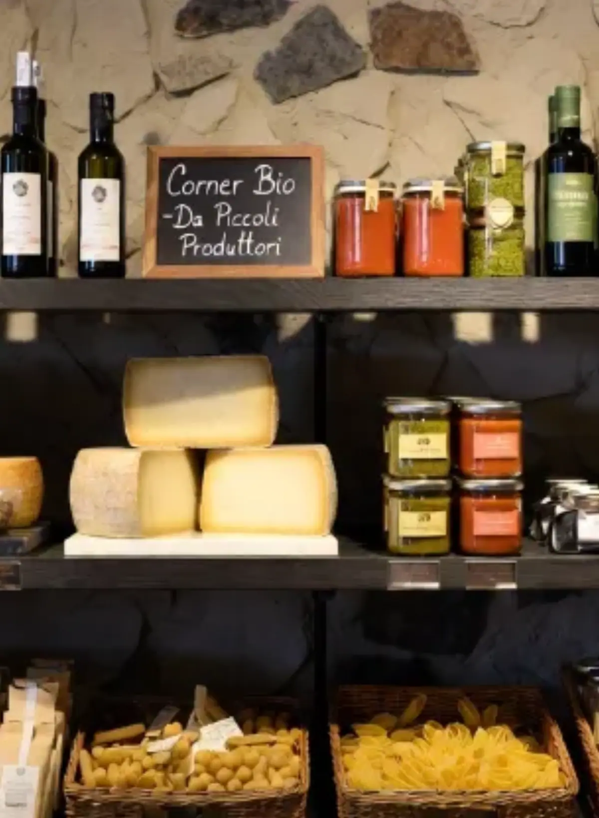 “Corner Bio” all’interno di un affascinante negozio gourmet italiano di alta gamma. Illuminazione naturale calda e morbida che crea un’atmosfera accogliente e di lusso. Bellissime mensole in legno scuro pregiato e parete in pietra naturale grezza. I prodotti biologici artigianali sono disposti con cura ed eleganza: eleganti bottiglie di olio extravergine di oliva (alcune beige, altre verdi), grandi forme di formaggi stagionati su piani di marmo bianco, barattoli di vetro con salse pregiate (pomodoro e pesto), vasetti di miele, prodotti al tartufo e pasta secca artigianale di lusso in cestini di vimini. Al centro un piccolo cartello in gesso dal gusto raffinato con la scritta “Corner Bio – Da Piccoli Produttori”.