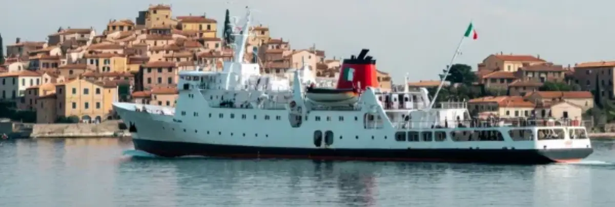 Una nave bianca più piccola (stile traghetto o yacht da crociera) si avvicina alla costa su acque tranquille e riflettenti. In secondo piano, su una collina nebbiosa e distante, un borgo medievale con mura di pietra, torri e case compatte color ocra e terracotta, circondato da vegetazione mediterranea e cipressi. Il cielo è velato da una leggera foschia che dà profondità e mistero, con luce morbida dell’alba o tramonto che sfuma i contorni del borgo. L’immagine evoca un senso di scoperta: la nave come ponte verso l’Italia autentica e nascosta.