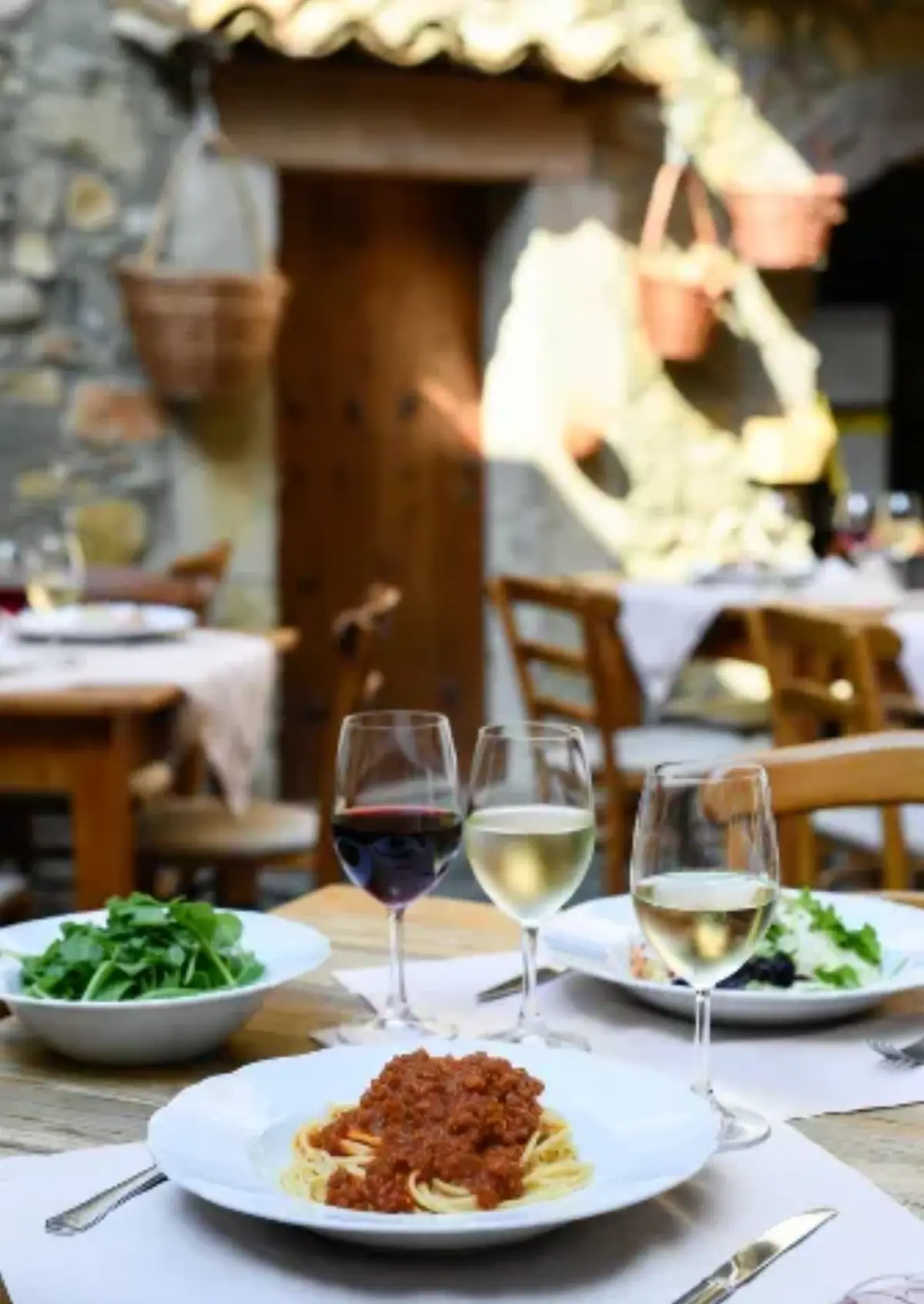 Interno realistico di una vecchia osteria in borgo di pietra. Tavolo di legno grezzo con tovaglia bianca semplice. Al centro un piatto di spaghetti al ragù con carne macinata e pomodoro, sopra una foglia di basilico fresco e un po’ di parmigiano grattugiato. Accanto: ciotola di insalata di rucola e pomodorini, due bicchieri di vino (uno rosso, uno bianco), forchetta e coltello posati, luce calda da una lampada appesa al soffitto con travi di legno scuro. Sfondo: muro in pietra grezza, porta antica socchiusa che dà su un vicolo, qualche bottiglia di vino sullo scaffale. Atmosfera intima, vissuta, niente turisti o roba patinata.