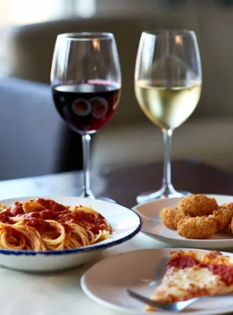un piatto fondo di spaghetti al pomodoro, con il sugo rosso vivo che ricopre generosamente la pasta, basilico fresco visibile e un aspetto invitante e fumante. Al centro-destra: una fetta di pizza margherita con pepperoni (o salame piccante), mozzarella filante e bordo croccante, servita su un piatto bianco con una forchetta accanto. Accanto alla pizza: un piccolo piatto con bastoncini di mozzarella fritta (o crocchette di formaggio), dorati e croccanti, che aggiungono un tocco di piatto veloce e sfizioso. Dietro i piatti, due calici di vino eleganti:Uno con vino rosso intenso e riflessi rubino. L'altro con vino bianco chiaro e luminoso.