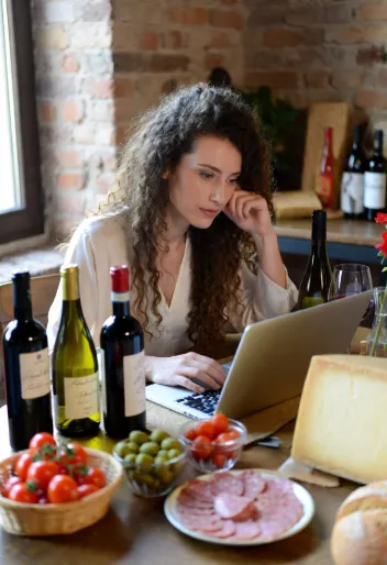 Una giovane donna con capelli ricci castani mossi, intorno ai 28-32 anni, aspetto naturale e autentico (tipo micro-influencer, non modella patinata). È seduta a un tavolo di legno rustico in una cucina/ sala da pranzo di campagna italiana con mattoni a vista. Indossa una camicia bianca morbida, leggermente sbottonata, maniche arrotolate. Davanti a lei un laptop aperto su cui sta scrivendo (probabilmente il guest post). Espressione concentrata ma serena, mano appoggiata al mento mentre guarda lo schermo. Intorno al tavolo, disposti in modo naturale e un po’ “instagrammabile ma non troppo”: bottiglie di vino rosso e bianco con etichette artigianali un cestino di vimini con pomodorini ciliegino freschi olive verdi in una ciotolina un pezzo di parmigiano stagionato pane casereccio un bicchiere di vino mezzo pieno Illuminazione calda, luce naturale da finestra laterale, atmosfera cozy e autentica da food blogger italiana.