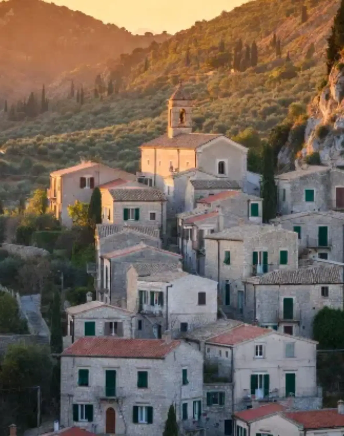 Un borgo italiano antico arroccato su una collina del Sud Italia (stile tipico di Puglia, Basilicata o Calabria) durante il tramonto dorato. Le case in pietra chiara e ocra sono strette le une alle altre, con tetti in tegole rosse antiche e persiane verdi. Vicoli stretti e tortuosi in ciottoli scendono verso la piccola piazza centrale dominata da un campanile in pietra.