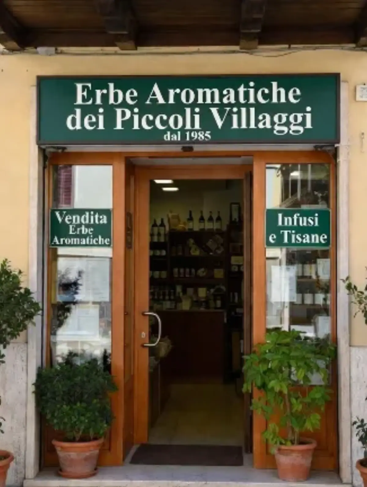 Una facciata rustica di un piccolo negozio in un paese di pietra, con intonaco chiaro e dettagli in legno antico. Sopra la porta d'ingresso in legno marrone con vetri a quadretti, c'è un'insegna rettangolare verde scuro con bordo decorato da foglie stilizzate in verde chiaro. Al centro dell'insegna, scritta in bianco maiuscolo elegante: Erbe Aromatiche dei Piccoli Villaggi