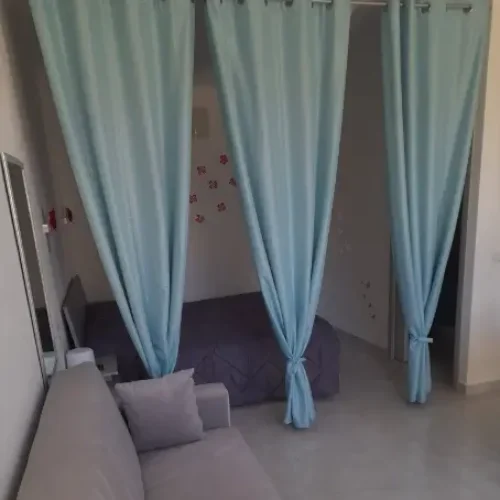Camera da letto piccola con letto matrimoniale nascosto dietro tende azzurre legate al centro, divano grigio in primo piano e pareti decorate con farfalle rosa.
