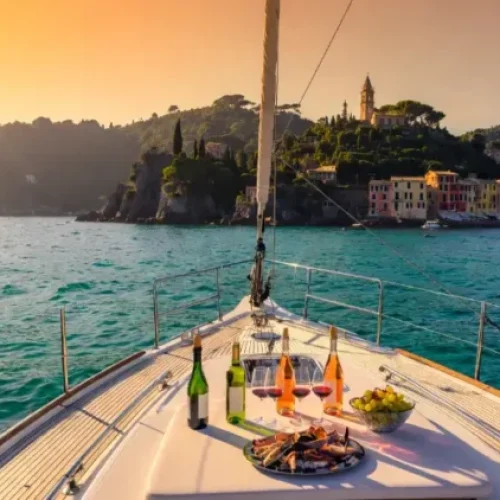 Yacht di lusso ancorato nella baia di Portofino al tramonto con bottiglie di vino bianco e rosé, calici e frutti di mare in coperta, vista sui colorati borghi liguri